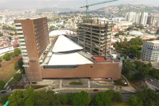 An aerial view of the Sentura Tlalnepantla Shopping Center under construction, showing a modern complex with a brown facade and triangular roof, surrounded by roads, trees, and the cityscape in the background.