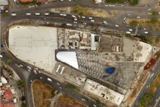 Aerial view of the Sentura Tlalnepantla Shopping Center, a large, partially constructed building with a triangular roof and exposed structure, surrounded by busy roads and vehicles in an urban area.