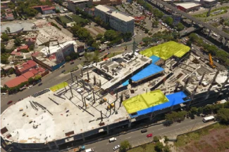Aerial view of the Sentura Tlalnepantla Shopping Center construction site in an urban area, with sections of the building highlighted in yellow and blue. The surrounding roads, vehicles, trees, and residential buildings are visible.