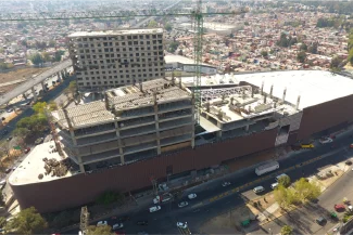 Aerial view of the Sentura Tlalnepantla Shopping Center under construction, with cranes, exposed steel beams, and construction materials. The site is surrounded by roads and cityscape, with cars and trucks visible on the streets.