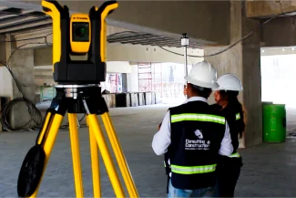 Two construction workers wearing safety vests and hard hats stand inside the unfinished Sentura Tlalnepantla Shopping Center, with a yellow measuring instrument on a tripod in the foreground.