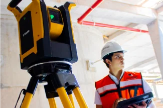 A construction worker wearing a hard hat and safety vest uses a tablet near a yellow measuring instrument on a tripod inside the Sentura Tlalnepantla Shopping Center, a building currently under construction.