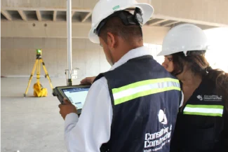 Two construction workers wearing reflective vests and white hard hats examine data on a tablet at the Sentura Tlalnepantla Shopping Center construction site; a yellow surveying tripod is in the background.