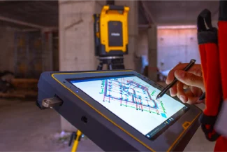 A person uses a stylus on a tablet displaying a construction plan at the Sentura Tlalnepantla Shopping Center construction site, with a surveying instrument in the background.