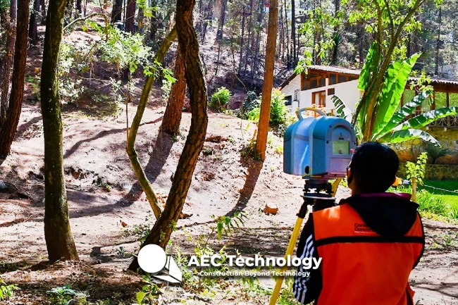 Una persona con chaleco de seguridad naranja inspecciona una zona boscosa con un escáner láser, realizando un levantamiento topográfico, con una casa visible entre los árboles al fondo. En la parte inferior aparece el texto "AEC Technology - Construction Beyond Technology".