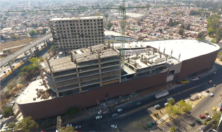 A large, partially constructed building with cranes on top rises near Sentura Shopping Center in Tlalnepantla, surrounded by busy streets and vehicles in a densely packed urban neighborhood of houses and roads