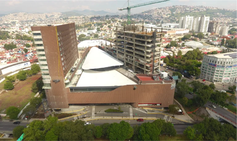Aerial view of the large, modern Sentura Shopping Center in Tlalnepantla, featuring a unique triangular roof beside an unfinished high-rise and crane. Roads, trees, and residential buildings dot the hilly landscape around the site