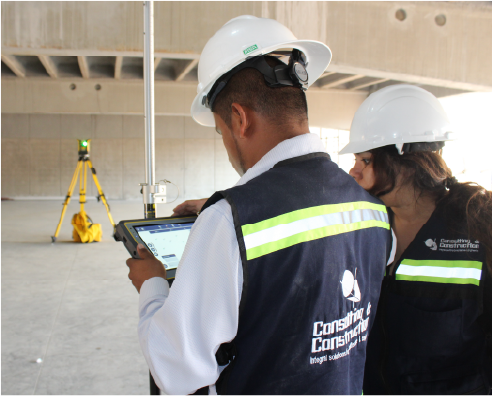 Dos obreros de la construcción con cascos y chalecos de seguridad revisan información en una tableta dentro de un edificio en construcción en el Centro Comercial Sentura. Al fondo se ve un trípode topográfico amarillo