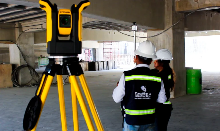Dos obreros de la construcción con chalecos de seguridad y cascos se encuentran en un edificio en construcción del Centro Comercial Sentura de Tlalnepantla. En primer plano, se ve un instrumento topográfico amarillo montado sobre un trípode entre el hormigón y los materiales de construcción