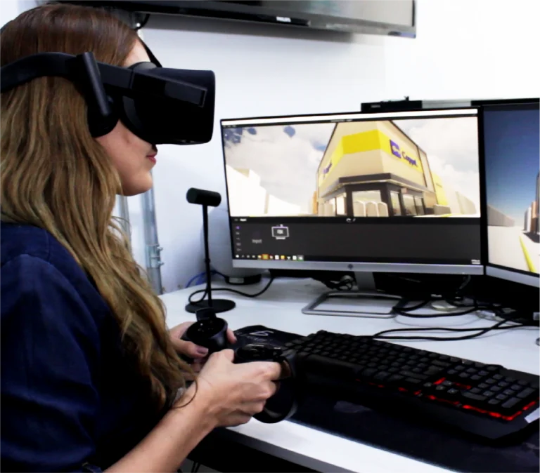 Una persona con un casco de realidad virtual utiliza unos controladores manuales mientras está sentada en un escritorio con un teclado y dos monitores que muestran un entorno de construcción virtual en 3D, posiblemente trabajando en un escaneo para Tienda Coppel Brisas