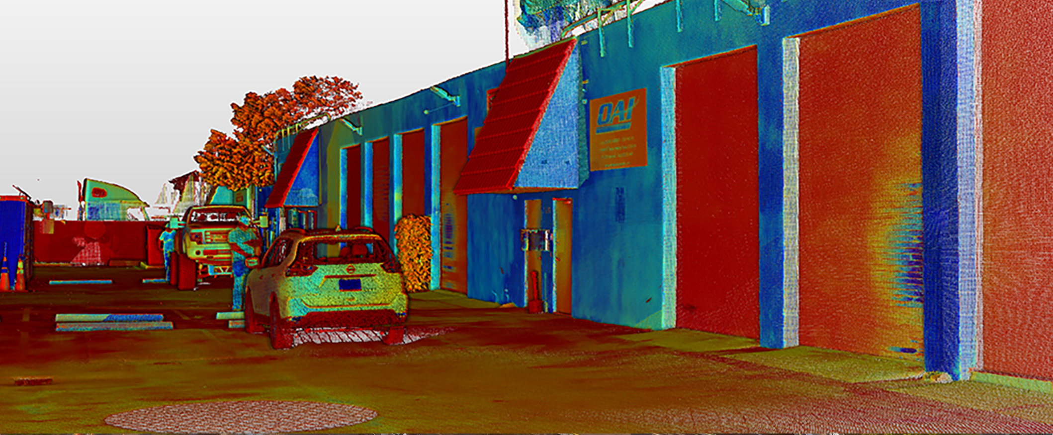 A colorized LiDAR scan shows a parking lot with cars and trucks parked by a large warehouse featuring multiple garage doors and red awnings. Overhaul Accessory equipment is visible near the building, with trees and fences in the background.