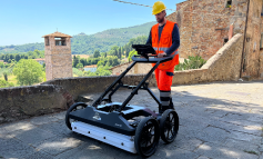 Un trabajador de la construcción con casco amarillo y chaleco de seguridad naranja opera un dispositivo de radar de penetración en el suelo en un camino de piedra al aire libre, mostrando captura de la realidad con edificios históricos y verdes colinas en el fondo.