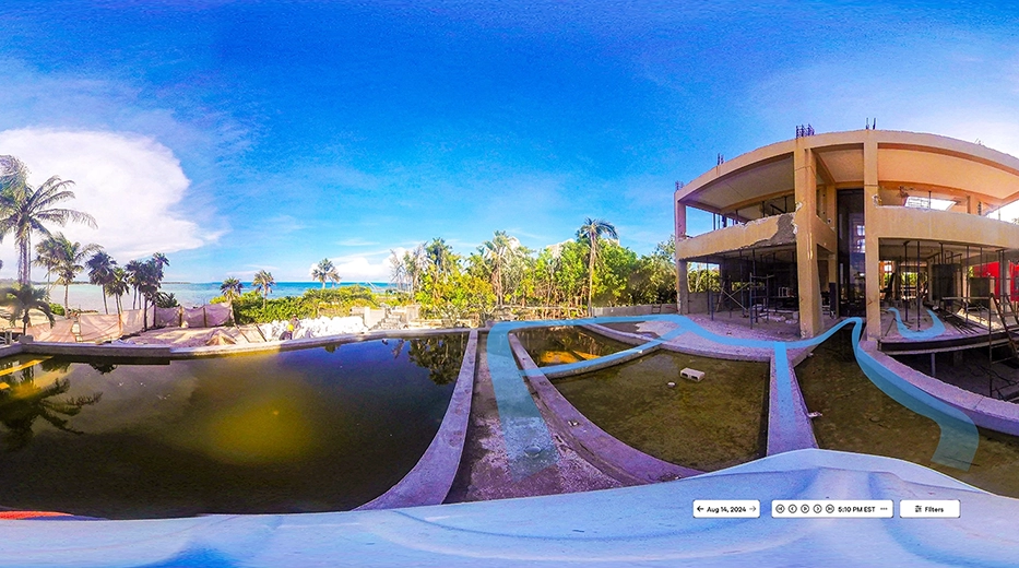 Una vista de 360 grados muestra un parque acuático abandonado con piscinas vacías o turbias, plantas crecidas y un gran edificio parcialmente abierto. Las palmeras y el océano aparecen al fondo bajo un cielo azul brillante, ideal para la fotografía 360 para la construcción.