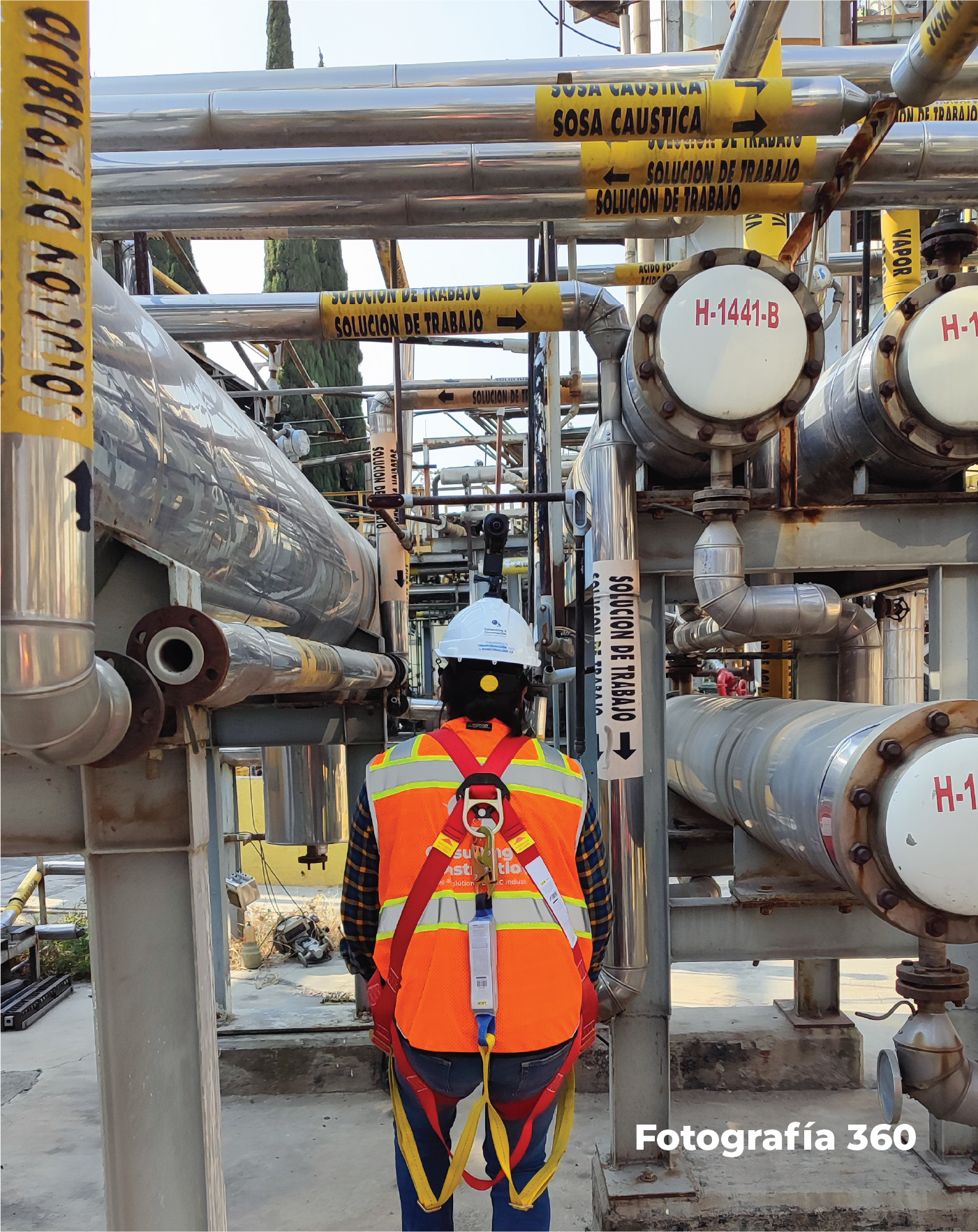 Un trabajador con chaleco de seguridad y casco se encuentra en una instalación industrial rodeado de tuberías y depósitos etiquetados con advertencias de seguridad, utilizando tecnologías avanzadas para documentar el entorno físico.