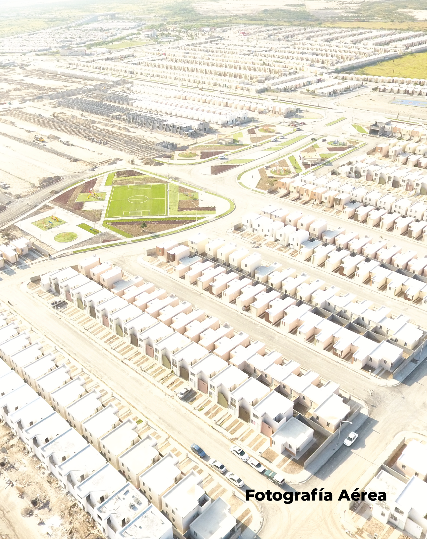 Vista aérea de un gran barrio residencial captada con tecnologías avanzadas, que muestra casas idénticas, un campo de fútbol verde, carreteras y coches. La zona está rodeada de terrenos sin urbanizar. El texto dice "Fotografía Aérea".