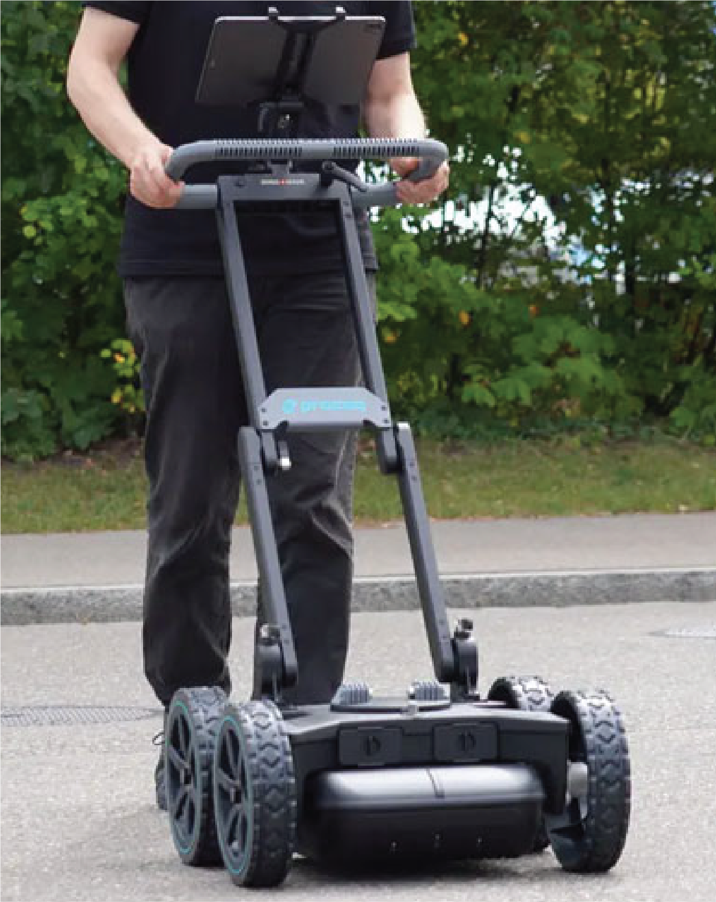 Utilizando tecnologías avanzadas, una persona opera un radar de penetración terrestre sobre ruedas, empujándolo por una superficie pavimentada al aire libre con vegetación de fondo para documentar el entorno físico.