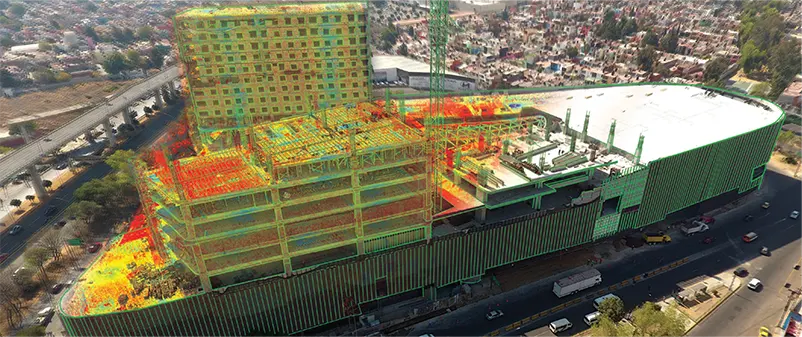 Vista aérea de una gran obra de construcción parcialmente escaneada en 3D utilizando datos de nubes de puntos de colores, superpuestos con líneas rojas, amarillas y verdes -como muestra de captura de la realidad y tecnologías avanzadas- rodeada de carreteras y zona urbana.