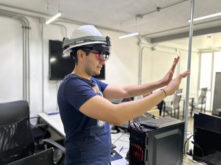 A person wearing a hard hat and augmented reality glasses interacts with extended reality content using hand gestures in an office with computer equipment and cables visible.