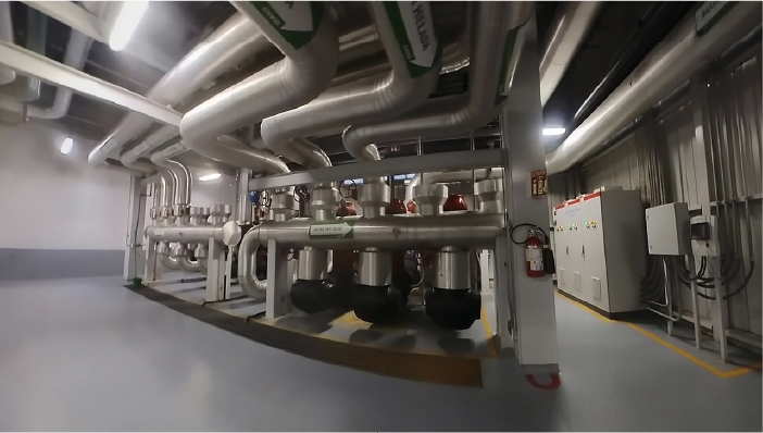 Una gran sala industrial con tuberías metálicas, conductos aislados, manómetros y paneles de control se captura mediante Fotografía Digital Técnica; un extintor está montado sobre un pilar, el suelo está limpio y la zona está bien iluminada.