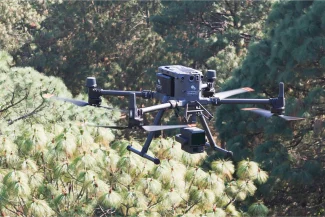 Un dron negro con cámaras y sensores sobrevuela los pinos de un bosque durante el día, captando la escena con precisión