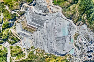 Fotografía Digital Técnica: Vista aérea de una gran cantera a cielo abierto rodeada de vegetación verde, con formaciones rocosas en terrazas, tierra expuesta y algunos edificios industriales en la parte inferior derecha.