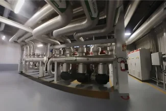 Una gran sala industrial con tuberías metálicas a lo largo del techo y las paredes, múltiples paneles de control y maquinaria aislada alineada en el suelo bajo una iluminación brillante, captada con Fotografía Digital Técnica.