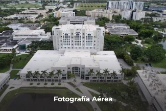 Vista aérea de un gran edificio blanco de varios pisos rodeado de palmeras y vegetación, con más edificios y un estanque en las inmediaciones; en la parte inferior aparece el texto Fotografía Digital Técnica con la leyenda Fotografía Aérea.