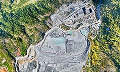 Vista aérea de una gran mina a cielo abierto rodeada de bosque verde, con tierra gris expuesta, carreteras construidas, balsas de agua y edificios industriales en la esquina superior derecha-una auténtica captura de la realidad industrial y natural.