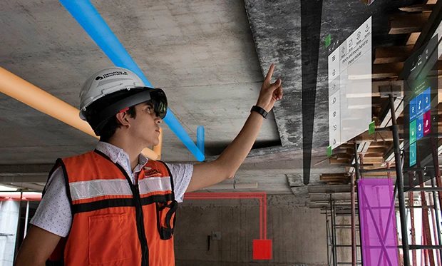 A construction worker wearing a hard hat and safety vest uses extended reality glasses, pointing at virtual interface panels projected onto a ceiling at a construction site with visible pipes and scaffolding.