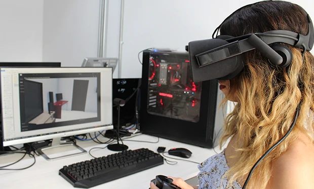 Una persona con un visor de realidad virtual y un control en la mano se sienta en un escritorio con una computadora, explorando la realidad extendida mientras observa un programa de modelado 3D en el monitor.