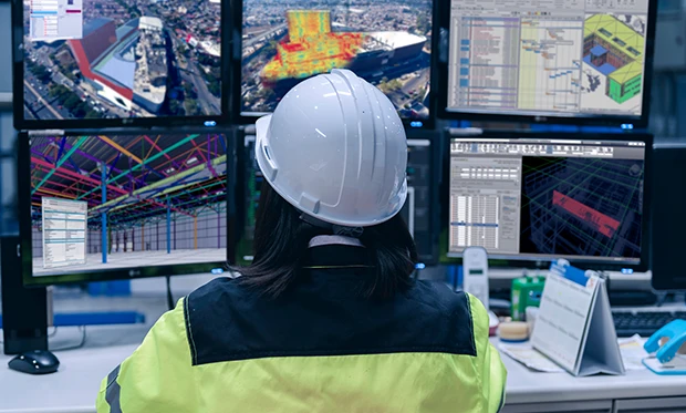 Una persona con casco blanco y chaqueta de alta visibilidad se sienta en un escritorio, utilizando tecnología en campo mientras observa planos de construcción, modelos digitales e imágenes aéreas en varios monitores de computadora.
