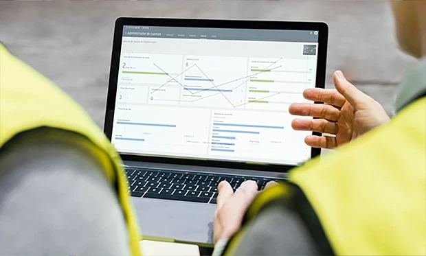 Dos personas con chalecos de seguridad amarillos revisan gráficos y diagramas en una computadora portátil, discutiendo análisis de datos y tecnología en campo en un entorno industrial activo.