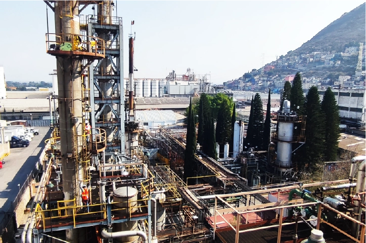 Una planta industrial con altas estructuras metálicas, tuberías y tanques se alza rodeada de árboles y edificios urbanos. La tecnología industrial LiDAR pudo cartografiar la zona, mientras una montaña se eleva al fondo bajo un cielo despejado.