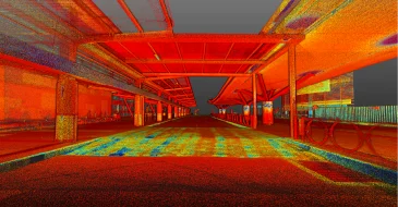 Colorida imagen de nube de puntos LiDAR de una estación de autobuses o terminal de tránsito vacía, con puntos rojos, naranjas, amarillos y azules que delinean la estructura, los andenes y el techo bajo un cielo oscuro.